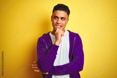 Young brazilian man wearing purple sweatshirt standing over isolated yellow background with hand on chin thinking about question, pensive expression. Smiling and thoughtful face. Doubt concept.