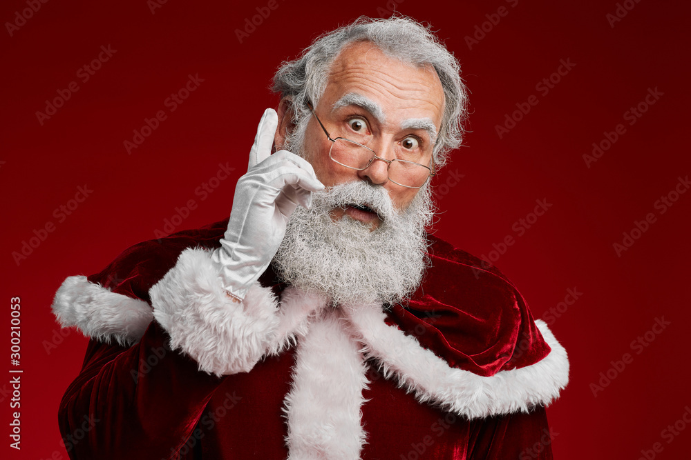 Portrait of surprised Santa Claus pointing up while posing against red background in studio, copy space