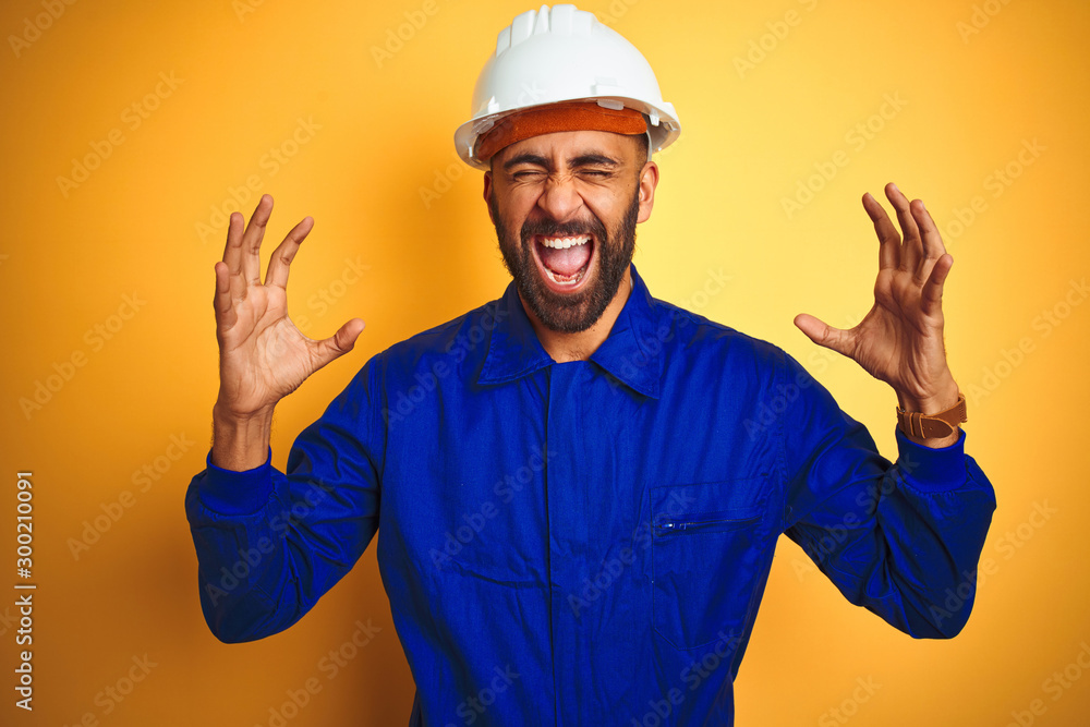 Handsome indian worker man wearing uniform and helmet over isolated yellow background celebrating mad and crazy for success with arms raised and closed eyes screaming excited. Winner concept Handsome indian worker man wearing uniform and helmet over isolated yellow background celebrating mad and crazy for success with arms raised and closed eyes screaming excited. Winner concept