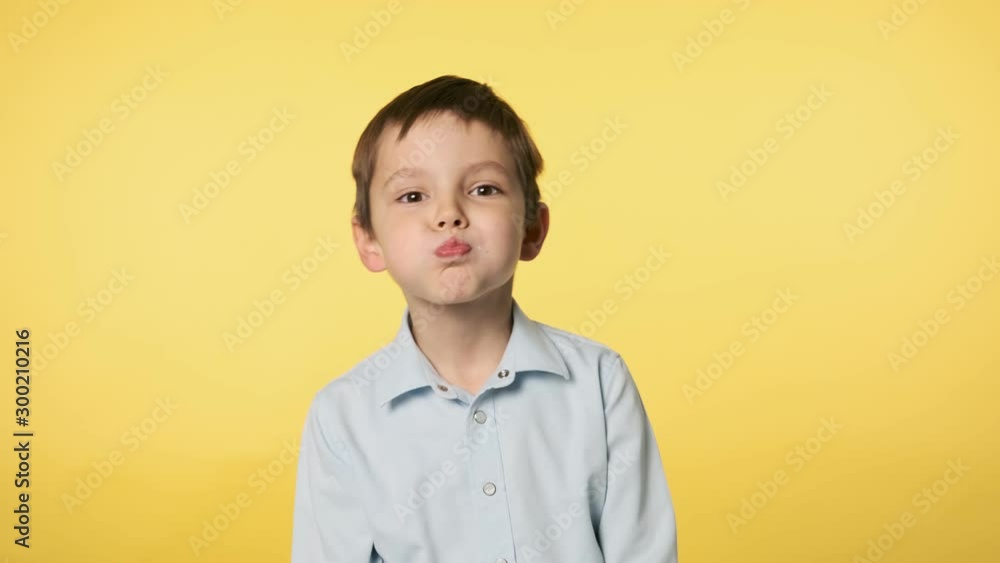 Funny little boy playfully making faces looking at camera. Positive emotion. Caucasian kid boy in blue shirt doing silly funny face puffing out cheeks at yellow background. Medium shot in 4k, UHD Funny little boy playfully making faces looking at camera. Positive emotion. Caucasian kid boy in blue shirt doing silly funny face puffing out cheeks at yellow background. Medium shot in 4k, UHD