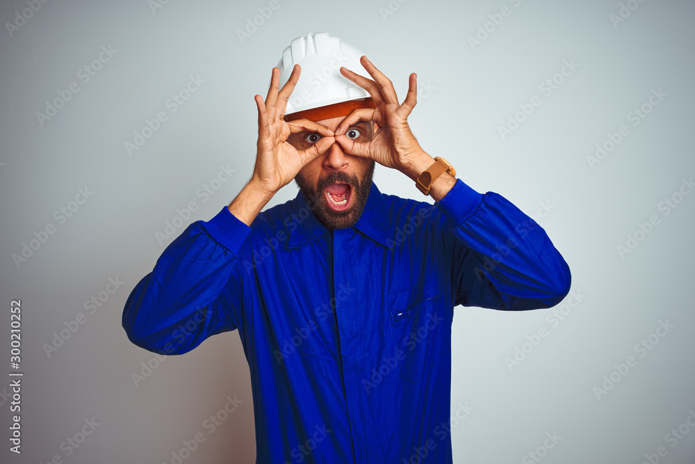 Handsome indian worker man wearing uniform and helmet over isolated white background doing ok gesture like binoculars sticking tongue out, eyes looking through fingers. Crazy expression. Handsome indian worker man wearing uniform and helmet over isolated white background doing ok gesture like binoculars sticking tongue out, eyes looking through fingers. Crazy expression.