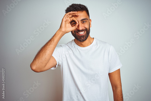 Young indian man wearing t-shirt standing over isolated white background doing ok gesture with hand smiling, eye looking through fingers with happy face. © Krakenimages.com