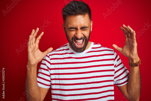 Young indian man wearing striped t-shirt standing over isolated red background celebrating mad and crazy for success with arms raised and closed eyes screaming excited. Winner concept