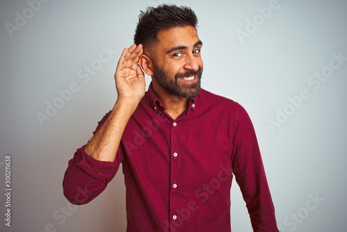 Young indian man wearing red elegant shirt standing over isolated grey background smiling with hand over ear listening an hearing to rumor or gossip. Deafness concept.