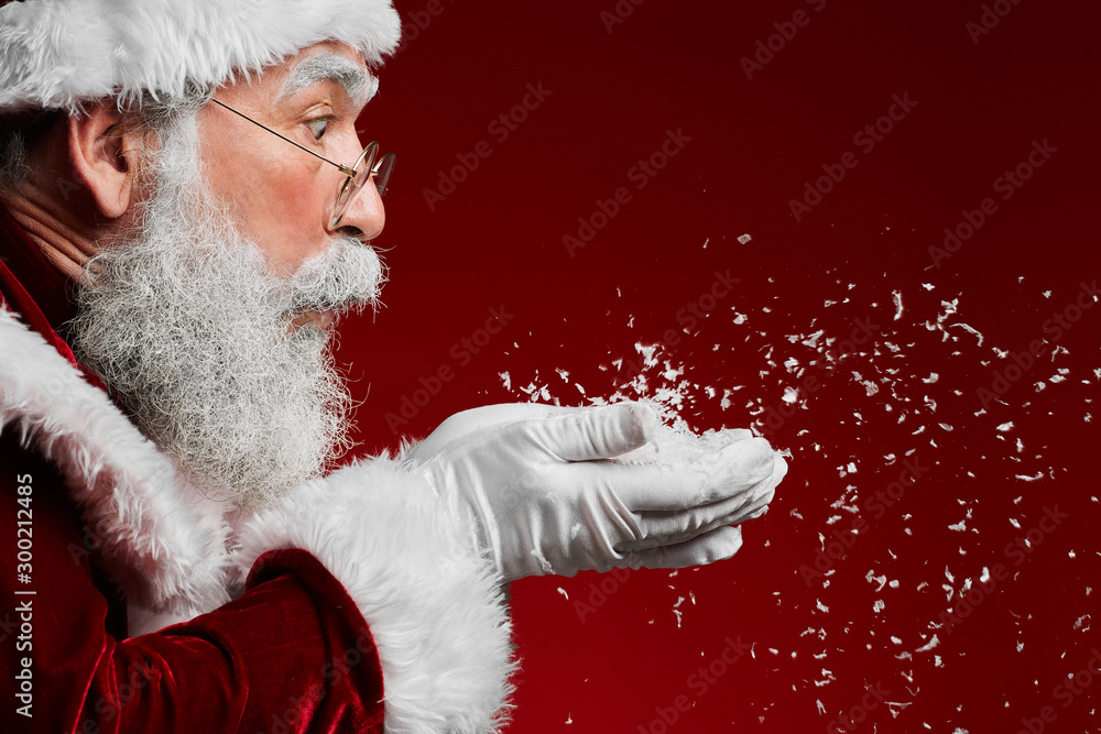 Side view portrait of classic Santa Claus blowing snow while standing against red background, copy space - obrazy, fototapety, plakaty 