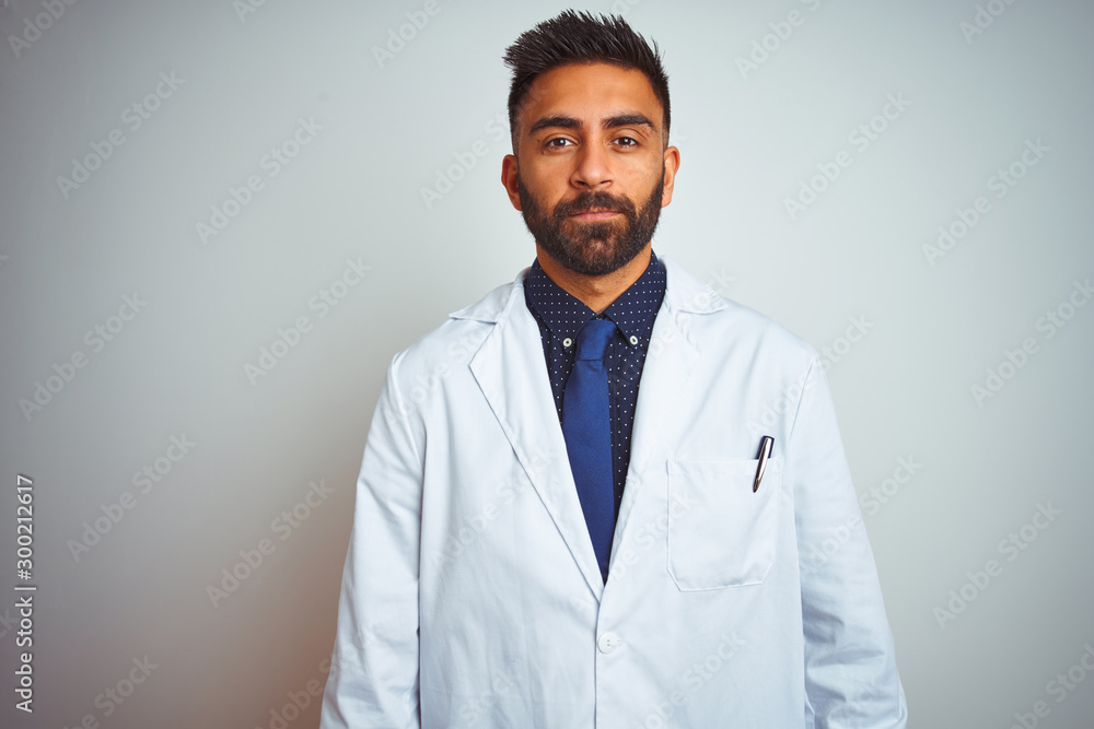 Young indian doctor man standing over isolated white background with serious expression on face. Simple and natural looking at the camera. Young indian doctor man standing over isolated white background with serious expression on face. Simple and natural looking at the camera.