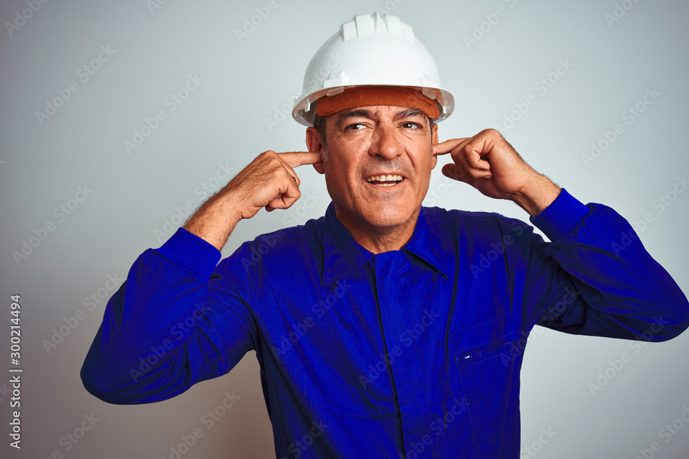 Handsome middle age worker man wearing uniform and helmet over isolated white background covering ears with fingers with annoyed expression for the noise of loud music. Deaf concept.
