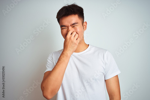 Young asian chinese man wearing t-shirt standing over isolated white background smelling something stinky and disgusting, intolerable smell, holding breath with fingers on nose. Bad smells concept.