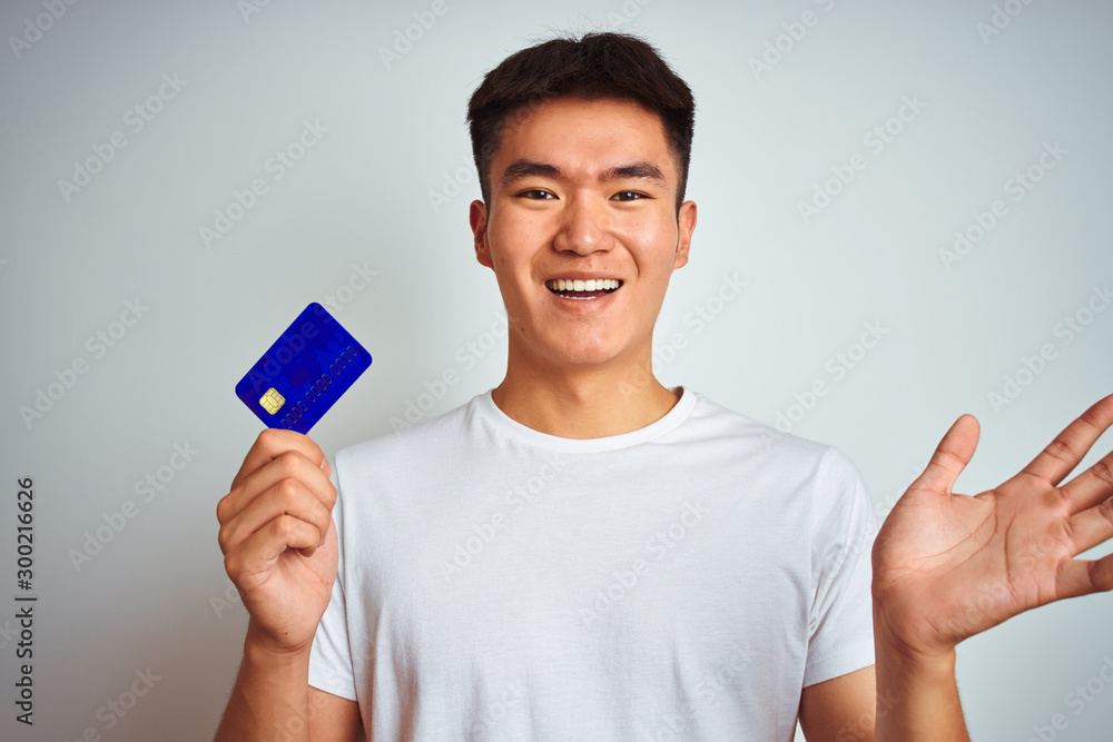 Young asian chinese man holding credit card standing over isolated white background very happy and excited, winner expression celebrating victory screaming with big smile and raised hands Young asian chinese man holding credit card standing over isolated white background very happy and excited, winner expression celebrating victory screaming with big smile and raised hands