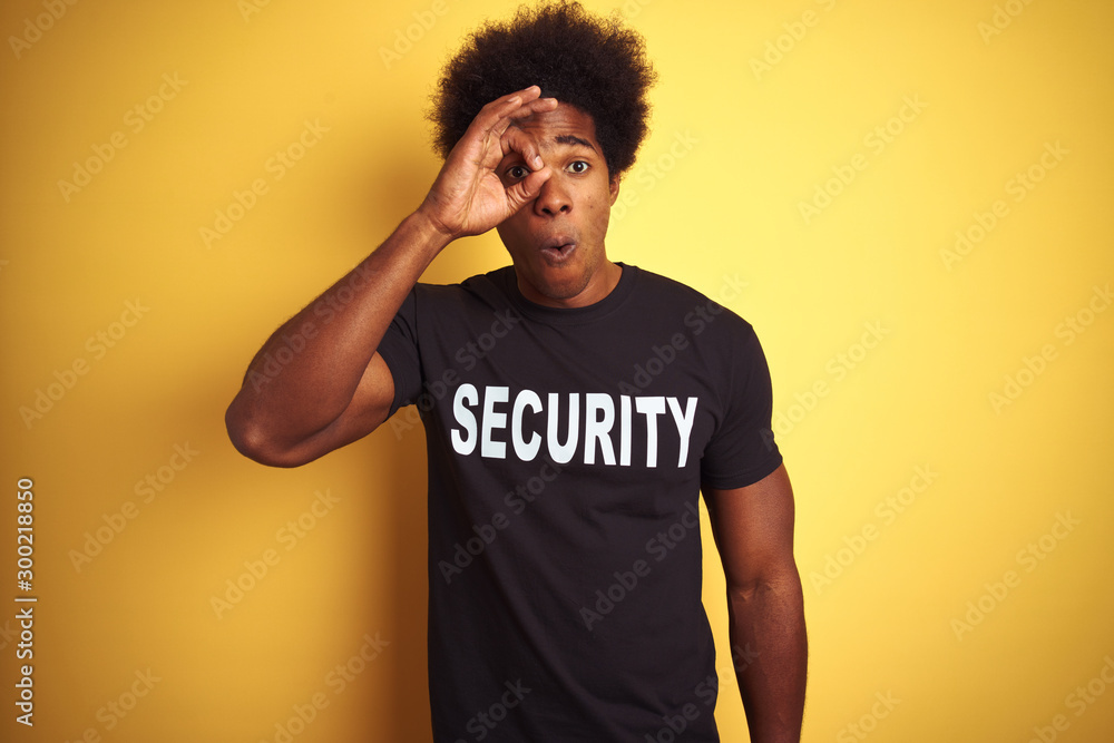 American safeguard man with afro hair wearing security uniform over isolated yellow background doing ok gesture shocked with surprised face, eye looking through fingers. Unbelieving expression. American safeguard man with afro hair wearing security uniform over isolated yellow background doing ok gesture shocked with surprised face, eye looking through fingers. Unbelieving expression.