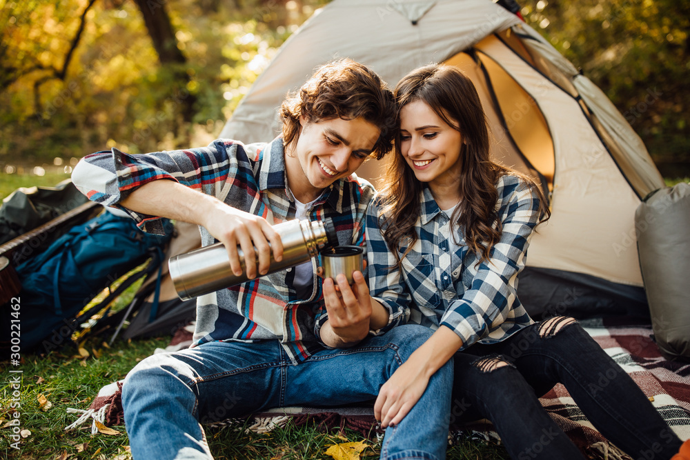 Young couple of tourists are exploring new places together. Attractive young woman and handsome man are spending time together on nature. Boyfriend pours tea from a thermos. Young couple of tourists are exploring new places together. Attractive young woman and handsome man are spending time together on nature. Boyfriend pours tea from a thermos.