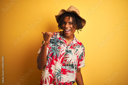 Afro american man with dreadlocks wearing floral shirt and hat over isolated yellow background smiling with happy face looking and pointing to the side with thumb up.