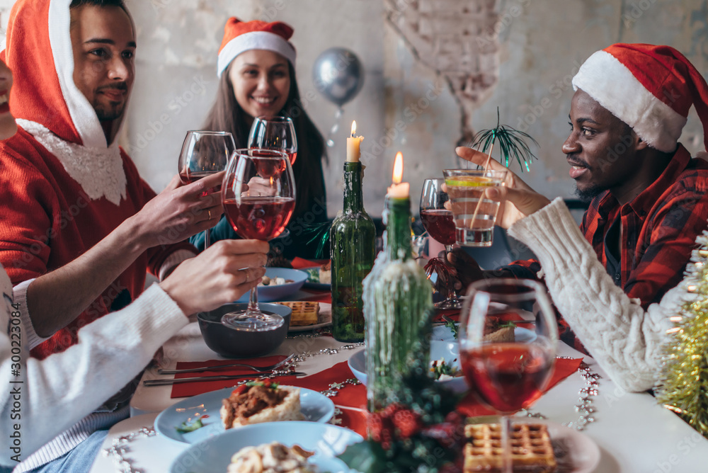People celebrating sitting at the table, clinking glasses. Christmas or New Year eve. - obrazy, fototapety, plakaty 
