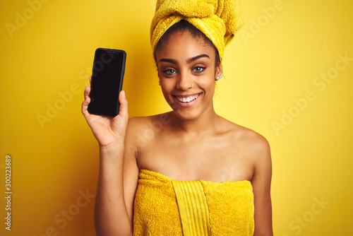 Afro woman wearing towel after shower showing smatrphone over isolated yellow background with a happy face standing and smiling with a confident smile showing teeth photo