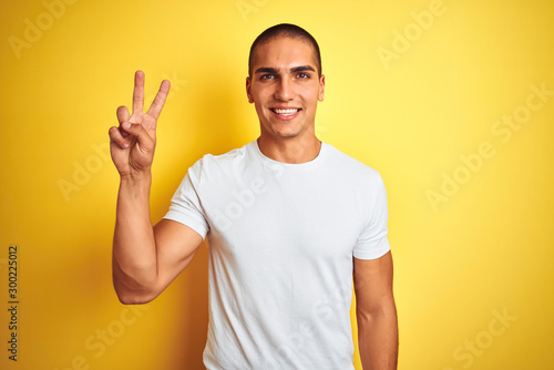Young caucasian man wearing casual white t-shirt over yellow isolated background smiling with happy face winking at the camera doing victory sign. Number two. © Krakenimages.com