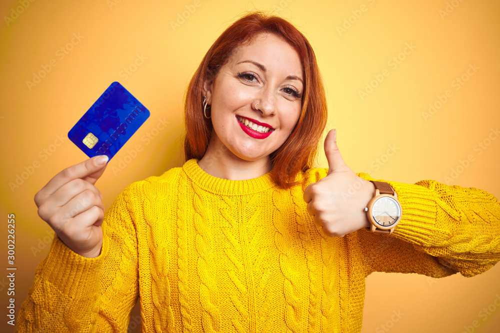 Young customer redhead woman holding credit card over yellow isolated background happy with big smile doing ok sign, thumb up with fingers, excellent sign Young customer redhead woman holding credit card over yellow isolated background happy with big smile doing ok sign, thumb up with fingers, excellent sign