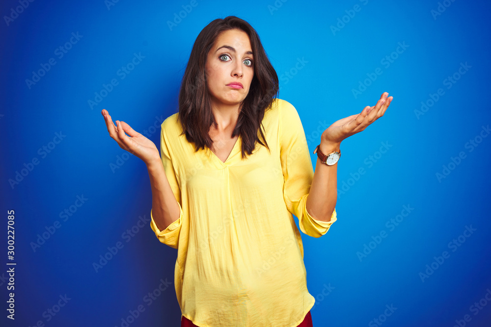 Young beautiful woman wearing yellow t-shirt standing over blue isolated background clueless and confused expression with arms and hands raised. Doubt concept. Young beautiful woman wearing yellow t-shirt standing over blue isolated background clueless and confused expression with arms and hands raised. Doubt concept.