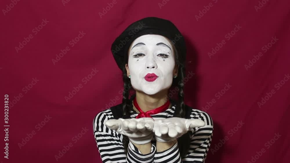 Beautiful girl mime sends air kiss and smiles looking at the camera. Beautiful girl in the image of a mime sends air kiss looking at the camera. Portrait on a red background. Beautiful girl mime sends air kiss and smiles looking at the camera. Beautiful girl in the image of a mime sends air kiss looking at the camera. Portrait on a red background.