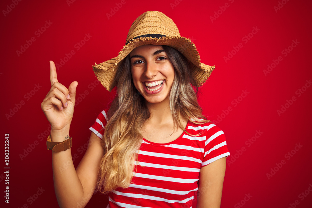Young beautiful woman wearing summer hat over red isolated background very happy pointing with hand and finger to the side Young beautiful woman wearing summer hat over red isolated background very happy pointing with hand and finger to the side