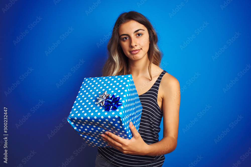 Young woman holding birthday present over blue isolated background with a confident expression on smart face thinking serious Young woman holding birthday present over blue isolated background with a confident expression on smart face thinking serious