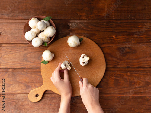 Female hands cut mushrooms. The concept of vegetarianism and a raw food diet. Champignon on a round wooden plate. Flat lay with copy space.