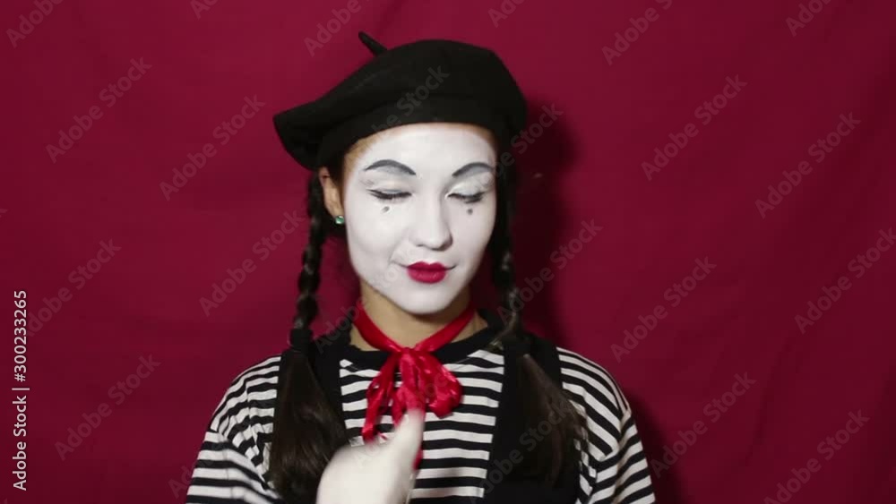 Beautiful girl mime smiles and winks eyes while looking at the camera. Beautiful girl in the image of mime winks and makes flirty movements with eyebrows looking at the camera. Portrait on a red backg Beautiful girl mime smiles and winks eyes while looking at the camera. Beautiful girl in the image of mime winks and makes flirty movements with eyebrows looking at the camera. Portrait on a red backg