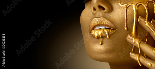 Gold Paint smudges drips from the face lips and hand, lipgloss dripping from sexy lips, golden liquid drops on beautiful model girl's mouth, gold metallic skin make-up. Beauty woman makeup close up