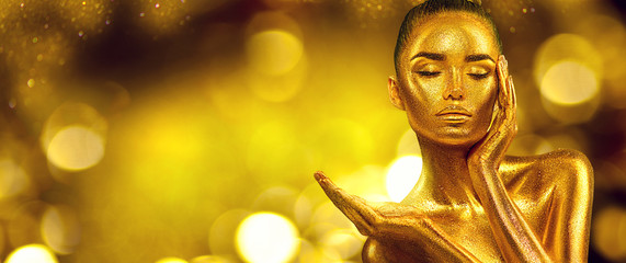 Gold Woman skin. Christmas Beauty fashion model girl with Golden make-up, hair and jewellery on gold background. Pointing hand, proposing products, sales. Metallic glance Fashion art portrait, glamour