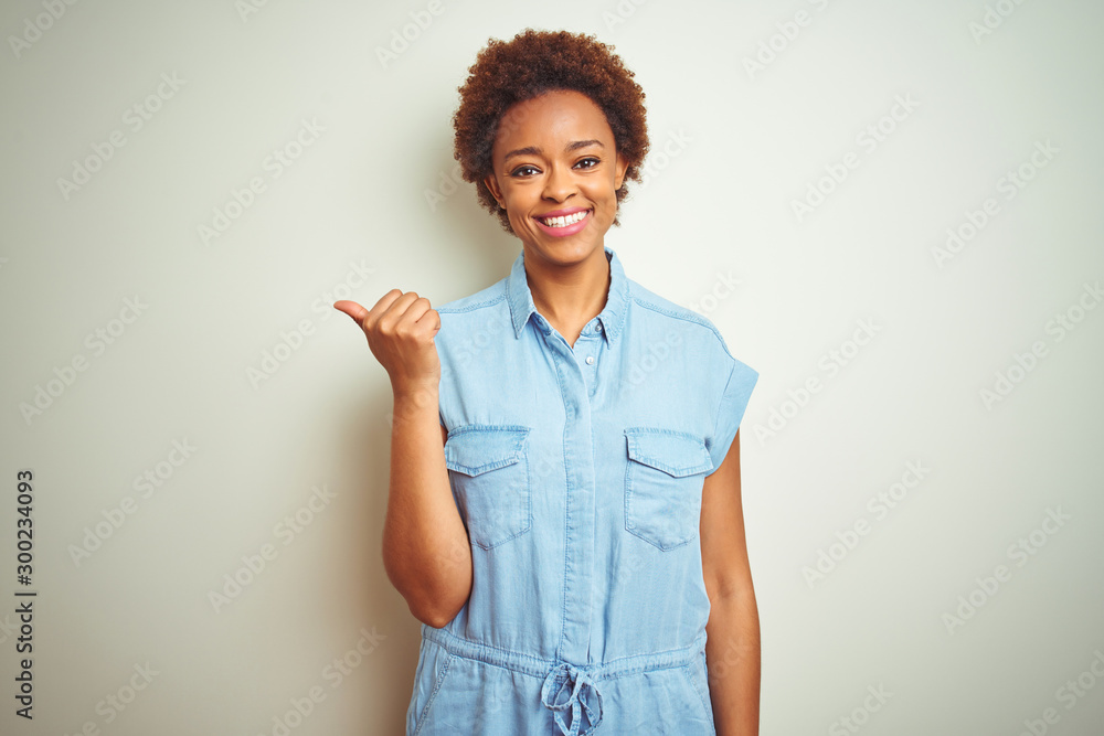 Young beautiful african american woman with afro hair over isolated background smiling with happy face looking and pointing to the side with thumb up. Young beautiful african american woman with afro hair over isolated background smiling with happy face looking and pointing to the side with thumb up.