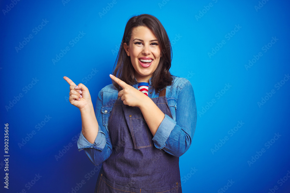 Fototapeta premium Young beautiful business woman wearing store uniform apron over blue isolated background smiling and looking at the camera pointing with two hands and fingers to the side.