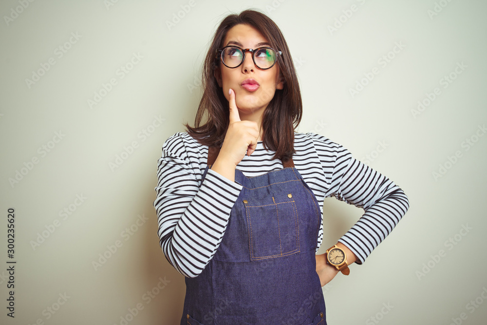 Young beautiful business woman wearing store uniform apron over isolated background with hand on chin thinking about question, pensive expression. Smiling with thoughtful face. Doubt concept. Young beautiful business woman wearing store uniform apron over isolated background with hand on chin thinking about question, pensive expression. Smiling with thoughtful face. Doubt concept.