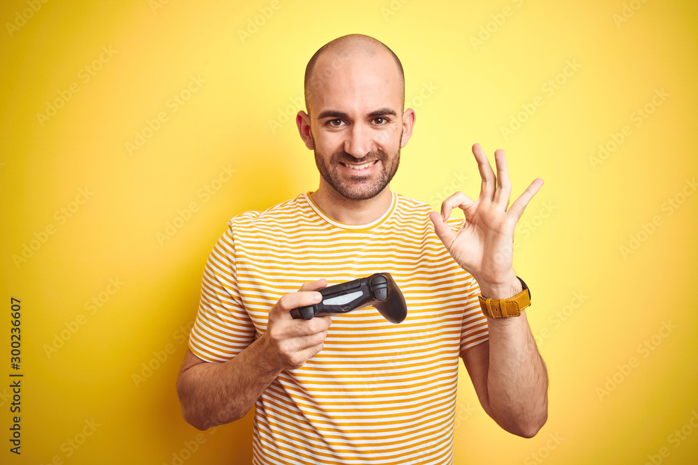Young man playing video games using joystick gamepad over isolated yellow background doing ok sign with fingers, excellent symbol Young man playing video games using joystick gamepad over isolated yellow background doing ok sign with fingers, excellent symbol