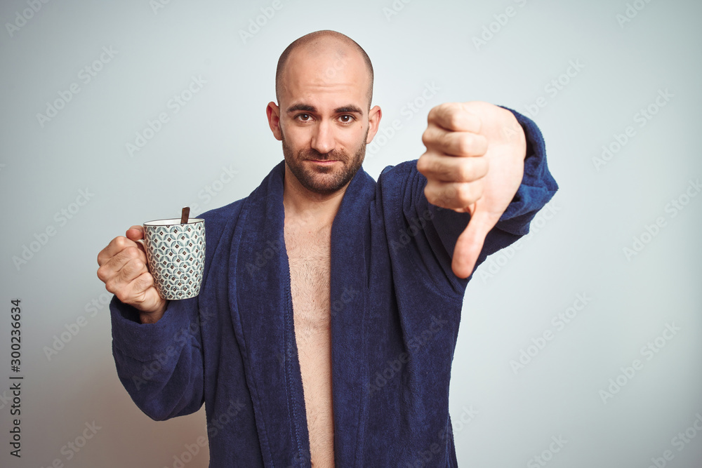 Naklejka premium Young man wearing a bathrobe drinking a cup of coffee in the morning over isolated background with angry face, negative sign showing dislike with thumbs down, rejection concept