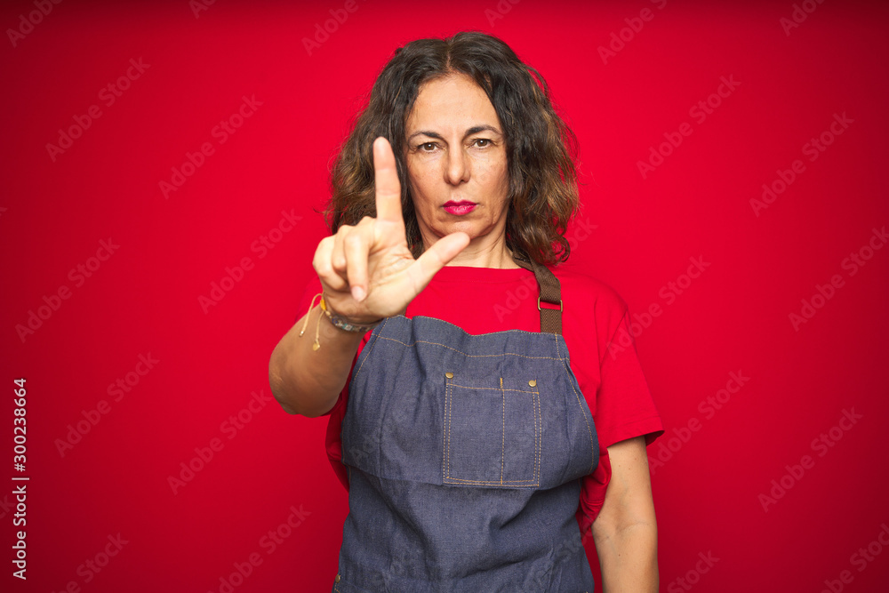 Middle age senior woman wearing apron uniform over red isolated background Pointing with finger up and angry expression, showing no gesture Middle age senior woman wearing apron uniform over red isolated background Pointing with finger up and angry expression, showing no gesture