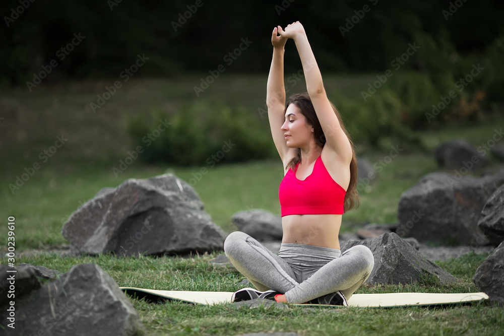 Fototapeta premium Young beautiful woman practicing yoga in the green park near stones. Wellness concept. Calmness and relax, woman happiness.