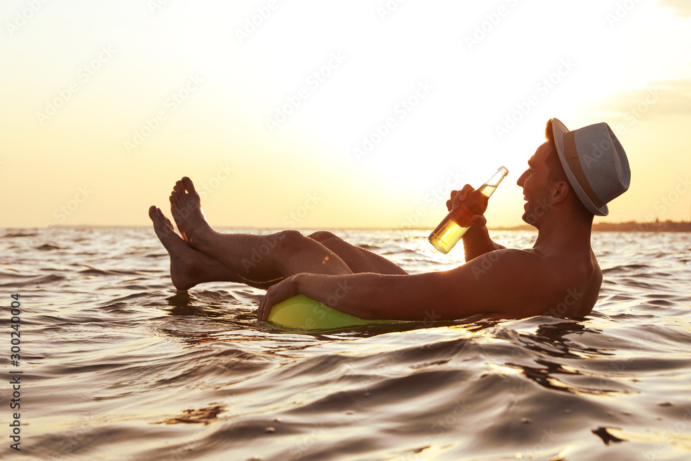 Young man with drink on inflatable ring in sea - obrazy, fototapety, plakaty 