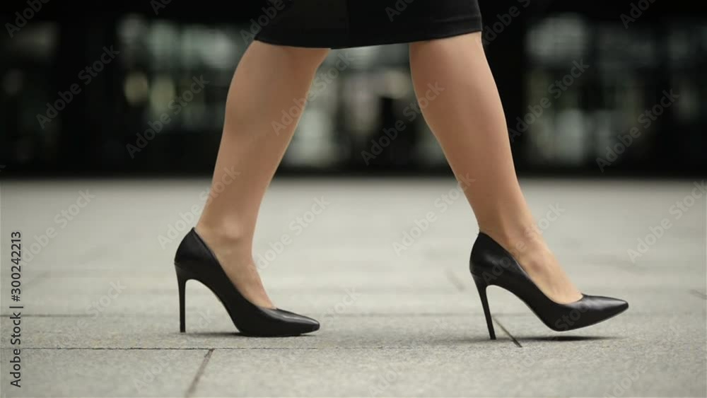 Feet Of Young Business Woman In High-Heeled Footwear Going In City. Businesswoman Legs In High-Heeled Shoes Walking Near Office After Work. Slow Motion. Feet Of Young Business Woman In High-Heeled Footwear Going In City. Businesswoman Legs In High-Heeled Shoes Walking Near Office After Work. Slow Motion.