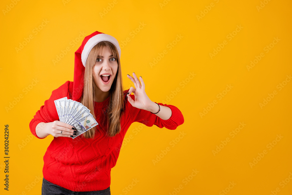 happy girl holding one hundred dollar bills in hand with ok gesture wearing santa hat isolated over yellow happy girl holding one hundred dollar bills in hand with ok gesture wearing santa hat isolated over yellow