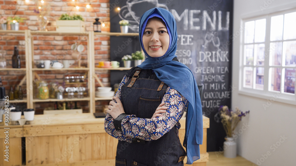 Arabian woman with happy smile wearing denim apron working in cafe bar. Islamic fashion lifestyle concept. charming young muslim lady crossed arms face camera with confident working in coffee shop
