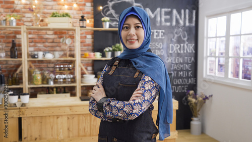 Arabian woman with happy smile wearing denim apron working in cafe bar. Islamic fashion lifestyle concept. charming young muslim lady crossed arms face camera with confident working in coffee shop