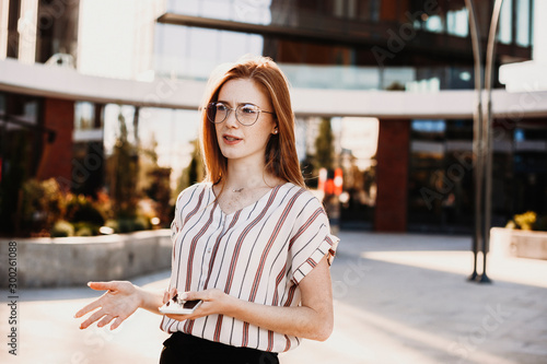 Lovely young female vlogger with red hair and freckles doing video content against a modern building. photo