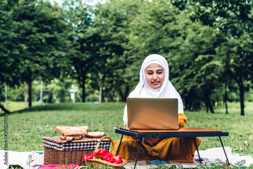 Portrait of happy arabic muslim woman with hijab dress smiling and using laptop cumputer in summer park photo