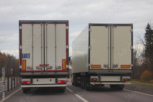 Two European white semi trucks with semi trailers overtaking on asphalt highway road on autumn day, rear view, transport logistics, goods delivery