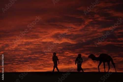 Silhouettes of two men with a camel (dromedary) in the desert against dark, red, cloudy sky.