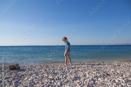 Cute European little boy in a blue swimming T-short is standing in the sea. He is enjoying his summer holidays. Back view.