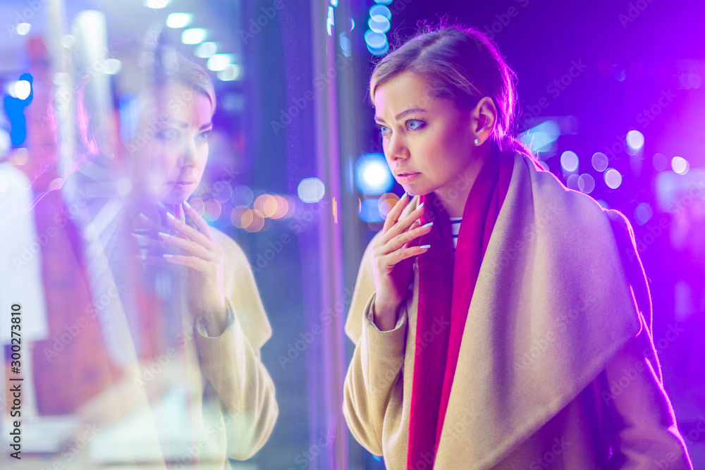 Christmas. A young beautiful woman in the late evening looks at the glowing window and chooses a gift. Colorful, magical lighting, beautiful glare. Cheerful girl wondered at the window, the agony of Christmas. A young beautiful woman in the late evening looks at the glowing window and chooses a gift. Colorful, magical lighting, beautiful glare. Cheerful girl wondered at the window, the agony of