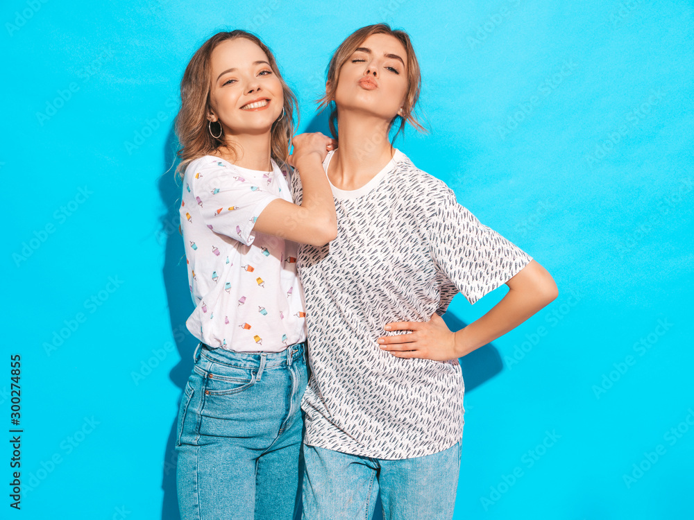 Two young beautiful smiling blond hipster girls in trendy summer colorful T-shirt clothes. Sexy carefree women posing near blue wall. Positive models having fun.Winks and makes duck face Two young beautiful smiling blond hipster girls in trendy summer colorful T-shirt clothes. Sexy carefree women posing near blue wall. Positive models having fun.Winks and makes duck face
