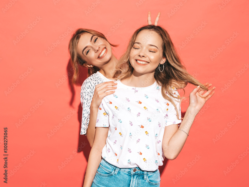 Two young beautiful smiling blond hipster girls in trendy summer colorful T-shirt clothes. Sexy carefree women posing near pink wall. Positive models having fun Two young beautiful smiling blond hipster girls in trendy summer colorful T-shirt clothes. Sexy carefree women posing near pink wall. Positive models having fun