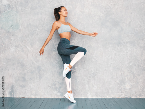 Portrait of fitness woman in sports clothing looking confident.Young female wearing sportswear.Beautiful model with perfect tanned body.Female jumping in studio near gray wall © halayalex