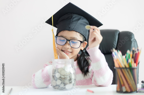 Little Asian girl wearing graduate hat putting the coin into clear glass jar piggy bank and smile with happiness for money saving to wealthness succesful in the future photo